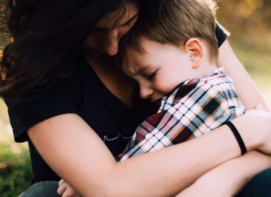 Woman sitting outdoors holding a young boy in her arms as he rests his head on her chest; both appear calm and close, sharing a quiet moment that helps regulate emotions.