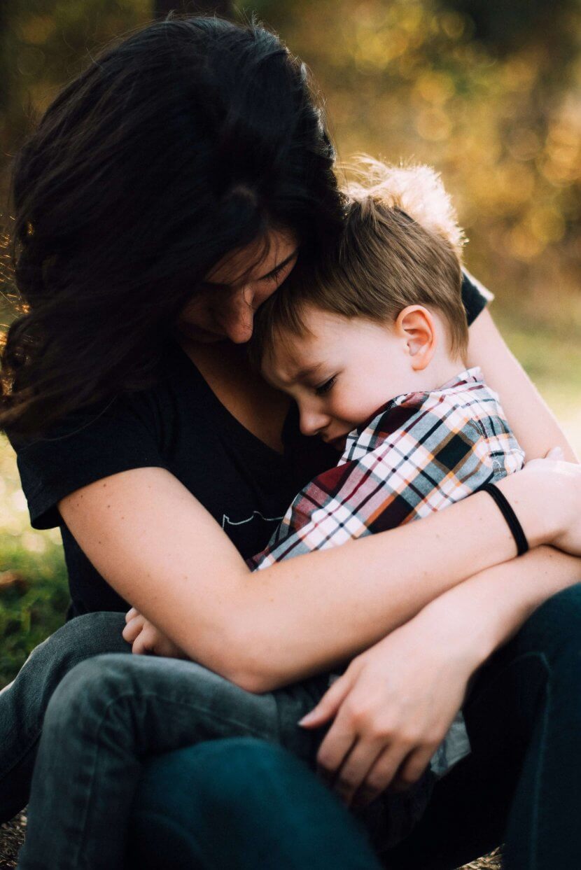 Woman sitting outdoors holding a young boy in her arms as he rests his head on her chest; both appear calm and close, sharing a quiet moment that helps regulate emotions.