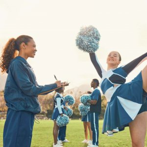 Cheerleading coach smiling and taking notes during practice with team on the field, representing supportive Sports Therapy in Scotch Plains, NJ with a trusted Sports Counselor in Scotch Plains, NJ.