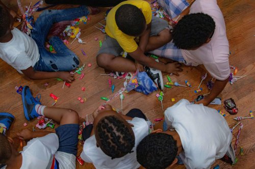 A group of children sit on a wooden floor, surrounded by scattered candy, selecting and sorting pieces.