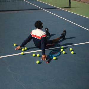 Athlete sitting on a tennis court surrounded by tennis balls and a racket, reflecting on challenges with performance. Represents support from a Sports Mental Health Therapist in Scotch Plains, NJ and Counseling for Athletes in Scotch Plains, NJ.