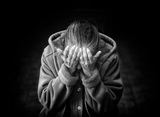 An elderly person sits with head bowed, covering their face with both hands, wearing a coat, against a dark background.