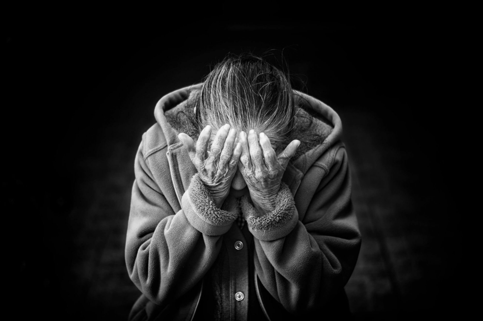 An elderly person sits with head bowed, covering their face with both hands, wearing a coat, against a dark background.