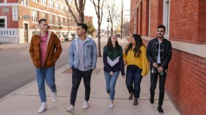Five young adults walk together on a city sidewalk next to a red brick building, talking and laughing on a clear day.