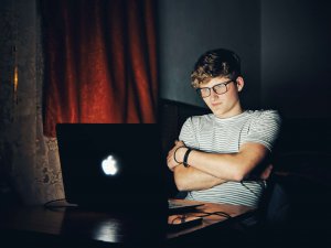 A person wearing glasses and a striped T-shirt sits with arms crossed in front of an open laptop in a dimly lit room.