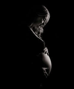 A pregnant woman stands in profile against a dark background, gently holding her belly and looking down.