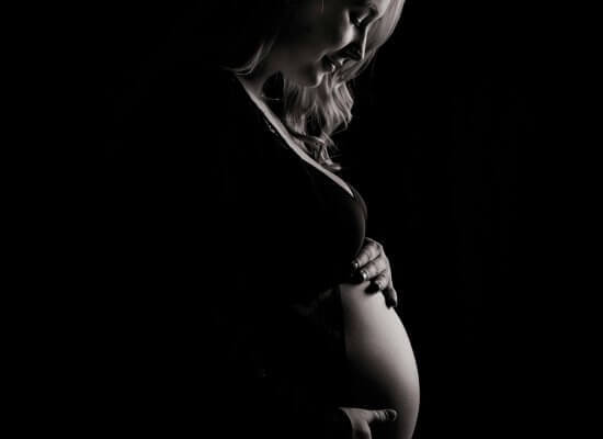 A pregnant woman stands in profile against a dark background, gently holding her belly and looking down.