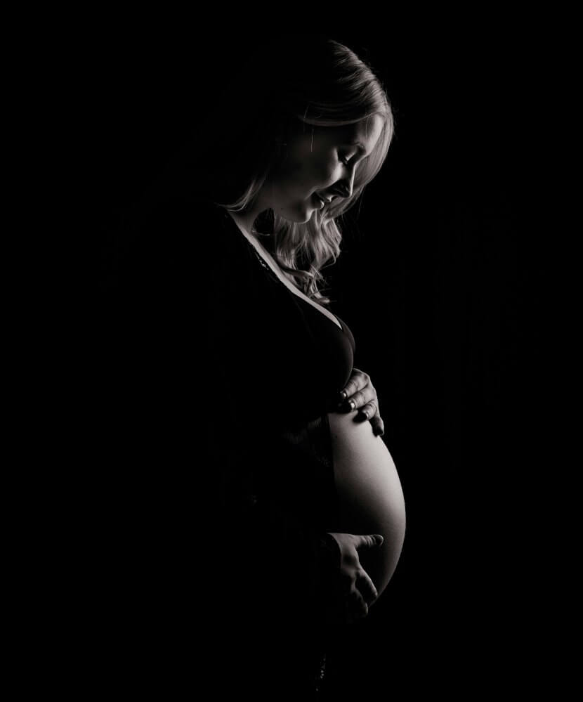 A pregnant woman stands in profile against a dark background, gently holding her belly and looking down.