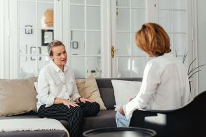 Two women sit facing each other in a bright room, engaged in conversation on a sofa and chair. One woman is talking while the other listens.