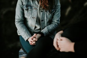 Two people are sitting close together, facing each other, with their hands clasped in their laps. One is wearing a denim jacket, and their faces are not visible.