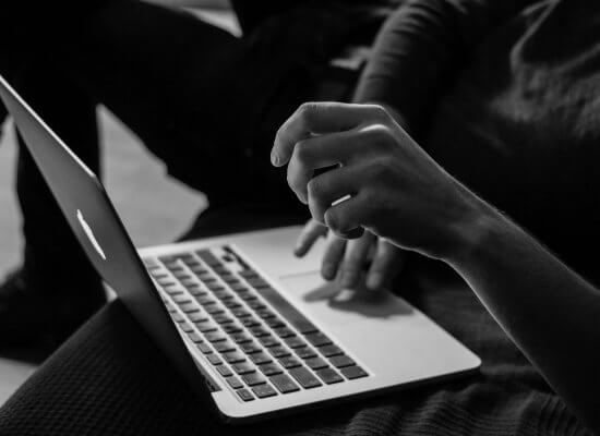 A person sits with a laptop on their lap, one hand on the keyboard and the other hand raised, in a grayscale setting.