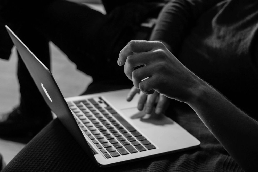 A person sits with a laptop on their lap, one hand on the keyboard and the other hand raised, in a grayscale setting.