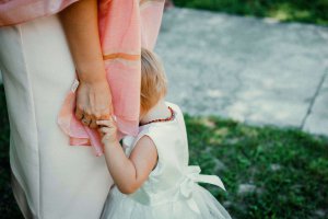 A small child in a white dress holds onto an adult’s hand and leg, partially hiding their face, while standing on grass next to a concrete path.