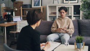 A young person sits on a couch talking to an adult with a clipboard, suggesting a therapy or counseling session in a modern office.