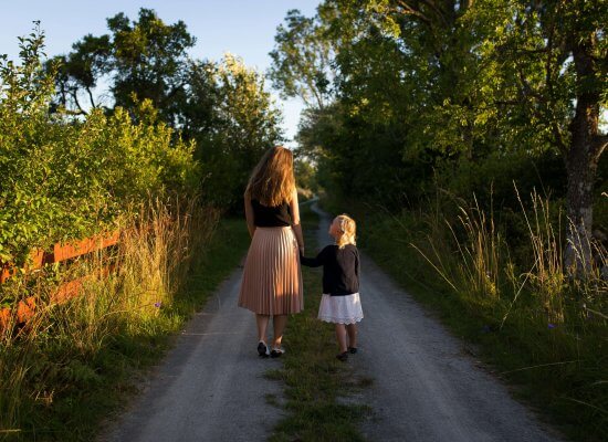 A woman and a young girl walk hand in hand down a tree-lined rural path in Branchburg NJ, with sunlight filtering through the trees.