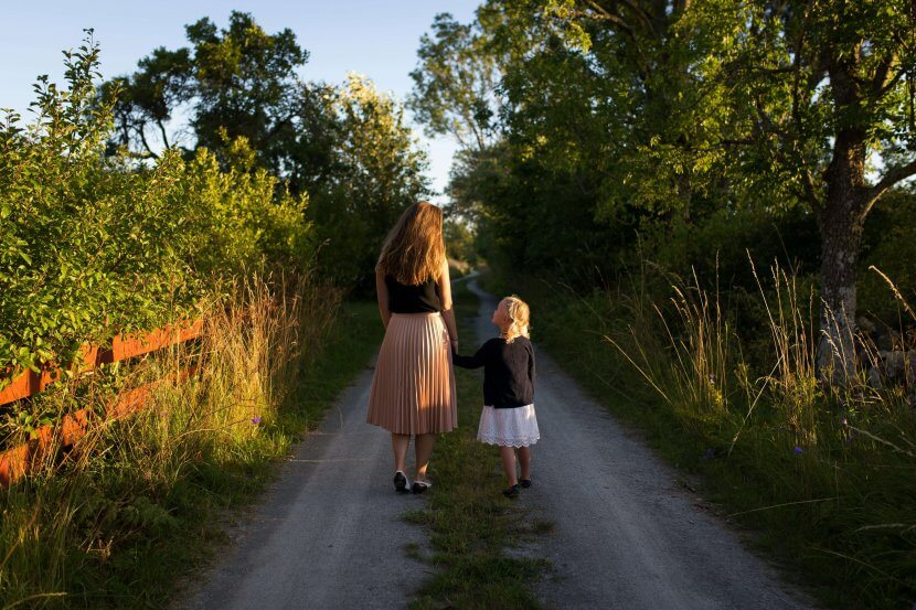 A woman and a young girl walk hand in hand down a tree-lined rural path in Branchburg NJ, with sunlight filtering through the trees.