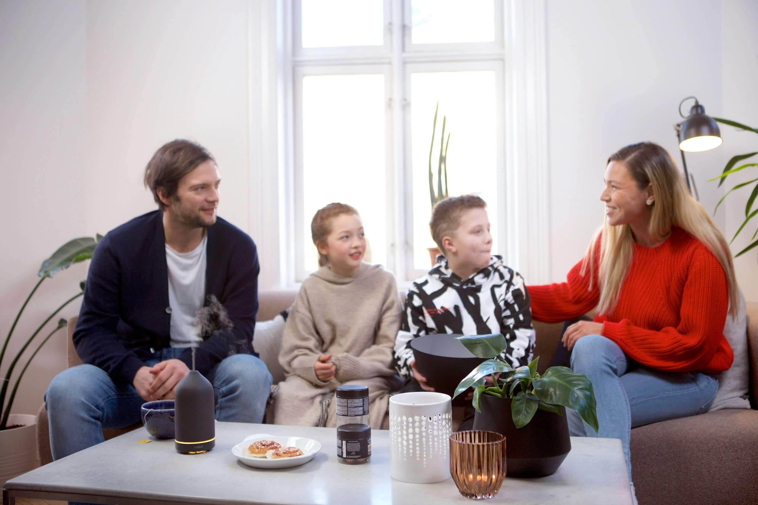 A warm family of four sits together in their bright, modern living room, demonstrating the positive family dynamics that result from effective parenting in Branchburg. This scene shows how NJ parents can create nurturing home environments where emotional regulation skills flourish. The comfortable, connected family interaction illustrates the benefits of emotional validation techniques that child therapists in Branchburg, NJ teach to strengthen family relationships.