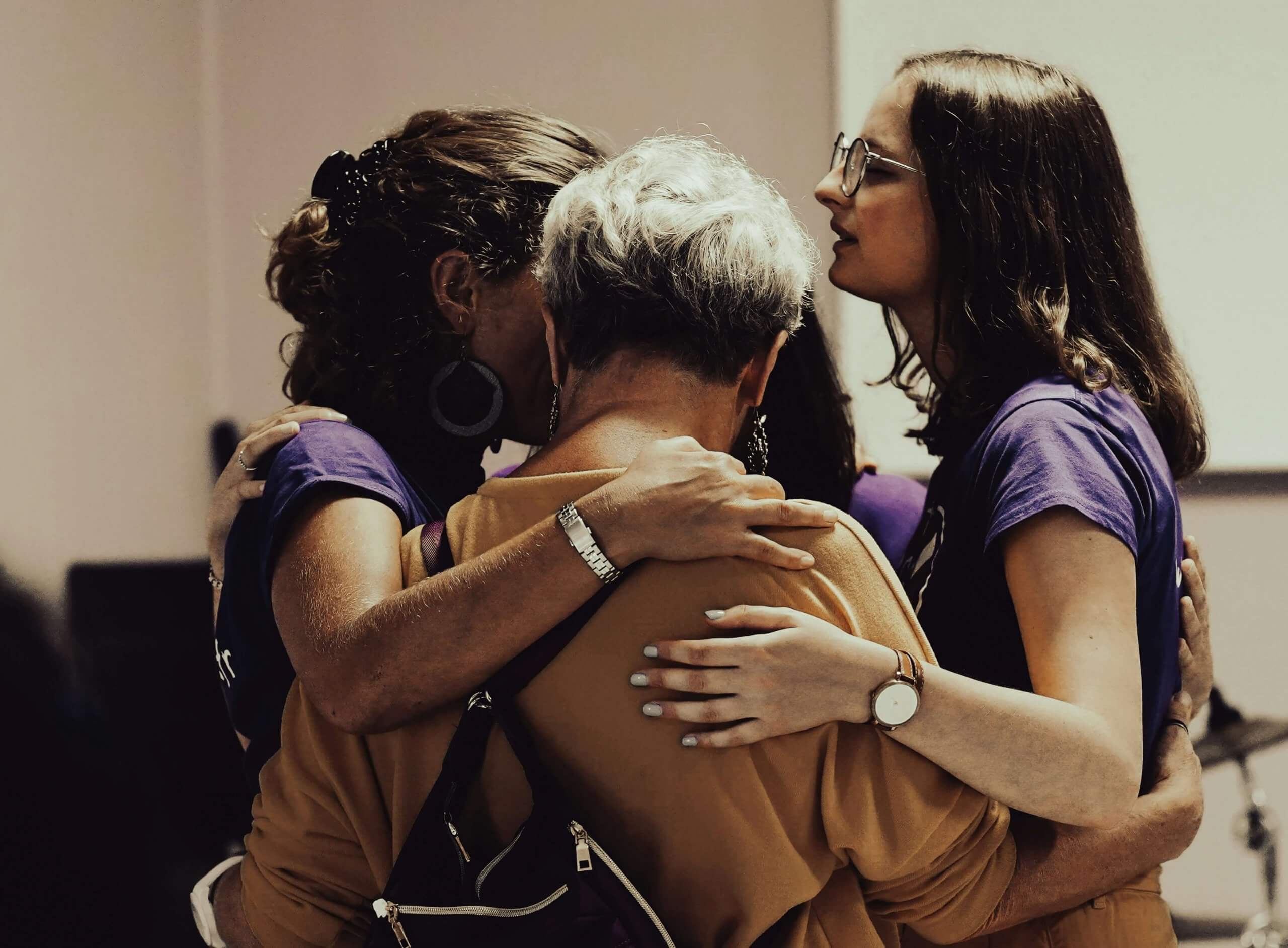 Four women in purple shirts embrace in a supportive group hug, representing the healing power of connection during grief. Child therapy and grief counseling services available in New Jersey, serving Scotch Plains 07076, Fanwood 07023, Garwood 07027, Clark 07066, and Westfield 07090.