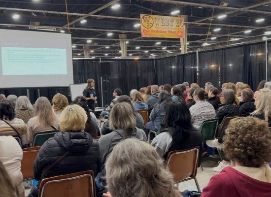 Lisa Weiss, LCSW, presents to a packed audience, speaking on the mental health of equestrians. She stands at the front of a large convention hall beside a projected presentation screen, facing rows of attendees seated in folding chairs. The crowd is engaged and attentive. A banner for the West 20 Horse Expo is visible overhead. Lisa Weiss specializes in therapy for equestrians, helping riders process traumatic falls, cope with the loss of a horse, and navigate barn conflicts — offering a safe, supportive space for healing rooted in an understanding of the horse world.