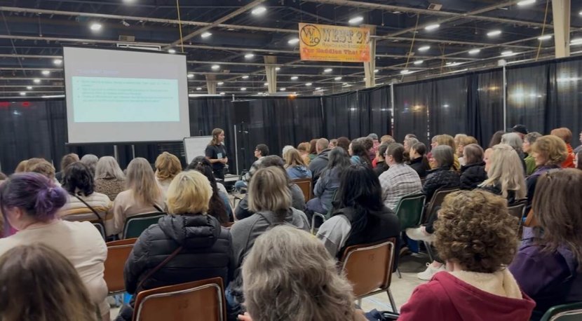 Lisa Weiss, LCSW, presents to a packed audience, speaking on the mental health of equestrians. She stands at the front of a large convention hall beside a projected presentation screen, facing rows of attendees seated in folding chairs. The crowd is engaged and attentive. A banner for the West 20 Horse Expo is visible overhead. Lisa Weiss specializes in therapy for equestrians, helping riders process traumatic falls, cope with the loss of a horse, and navigate barn conflicts — offering a safe, supportive space for healing rooted in an understanding of the horse world.