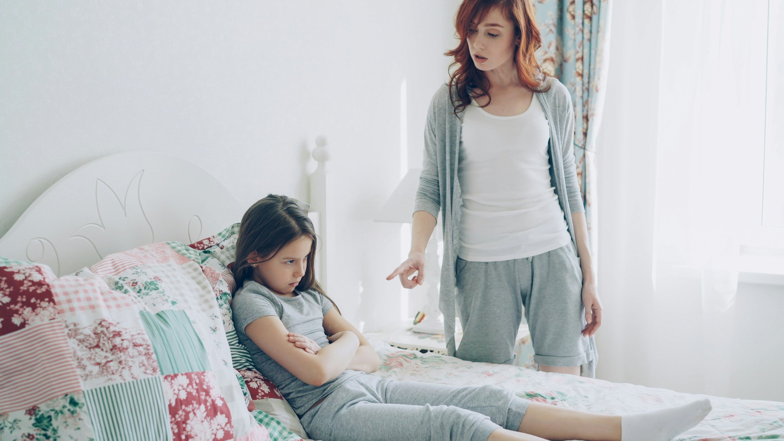 A frustrated girl sitting on her bed with arms crossed while her mother tries to talk to her, illustrating the challenge of explaining therapy to a child in Scotch Plains, NJ. Serving families in Branchburg, Somerville, and surrounding New Jersey communities seeking parenting advice and parent coaching in Scotch Plains, NJ.