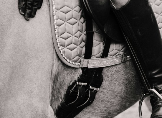 Close-up black and white photograph of quilted white saddle pad, black leather saddle, riding gloves, and equestrian equipment on horse, showing professional riding gear central to equestrian-related trauma therapy.