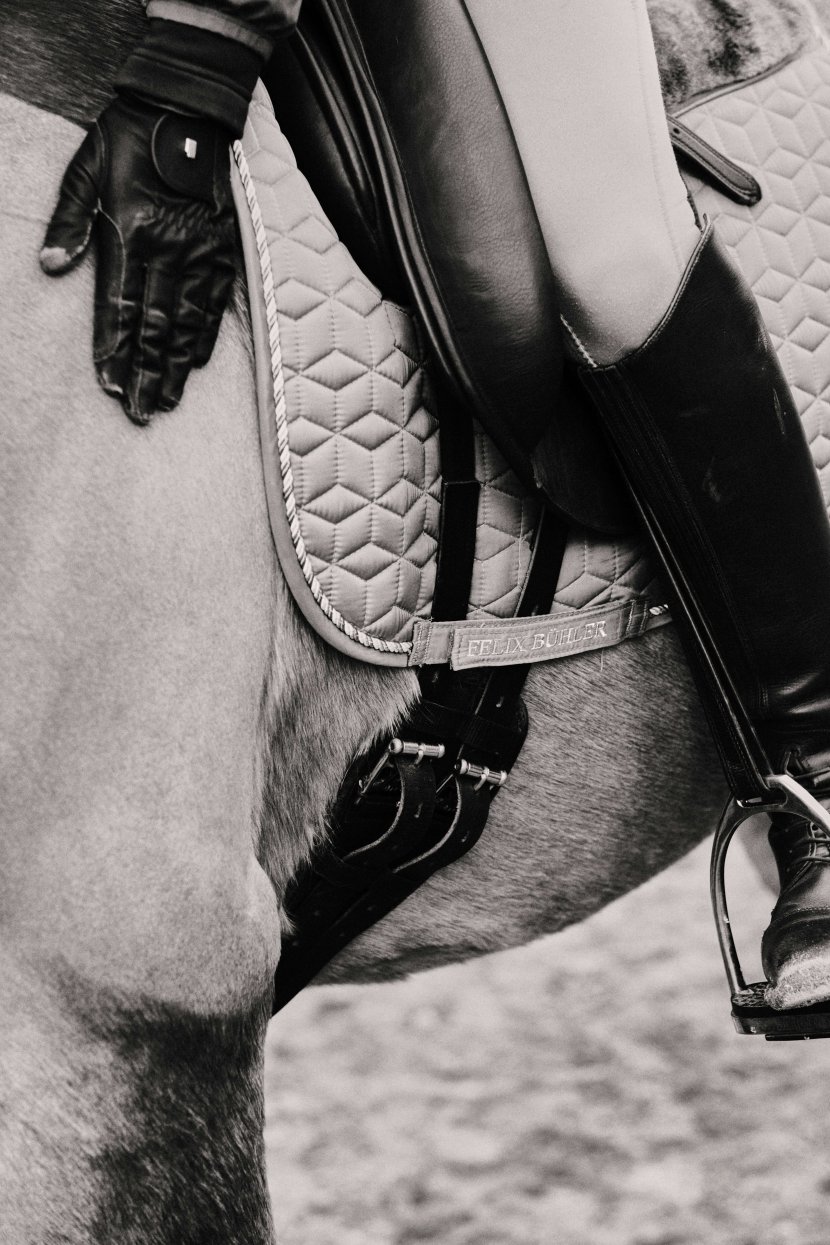 Close-up black and white photograph of quilted white saddle pad, black leather saddle, riding gloves, and equestrian equipment on horse, showing professional riding gear central to equestrian-related trauma therapy.