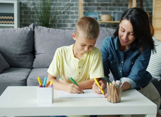 A smiling mother sits beside her son as he draws with colorful markers at a white table, capturing the essence of emotional wellness NJ in Somerville. Serving Somerville, Branchburg, and Scotch Plains — providing emotional regulation skills NJ families can use to nurture emotional growth NJ children deserve.