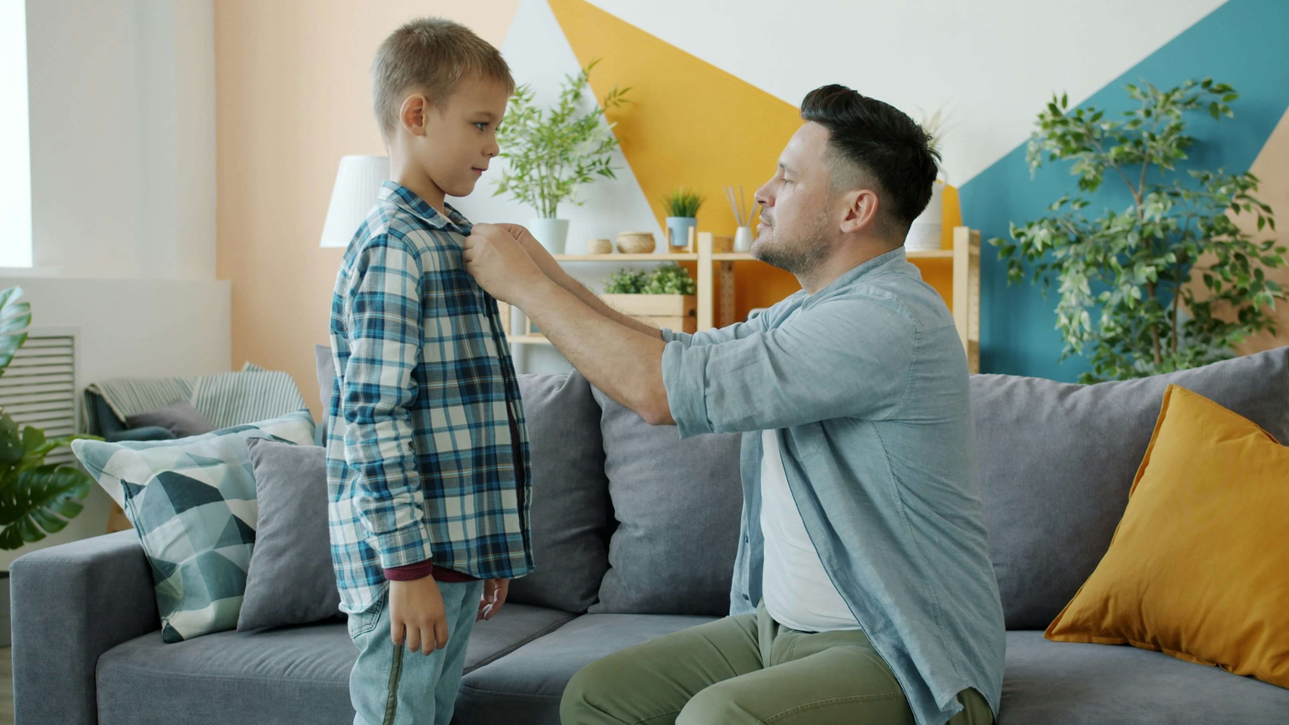 A caring father kneels to help his young son button his shirt in a warm living room, reflecting mindful parenting NJ values in Branchburg. Serving Branchburg, Somerville, and Scotch Plains — offering emotional validation NJ parents and children need to build secure, trusting relationships.